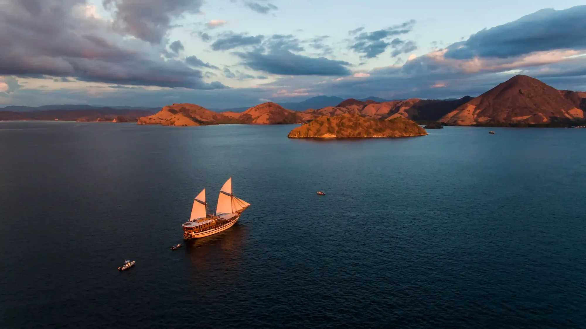Aerial view of a phinisi with tenders on a calm bay below hills lit by low golden light